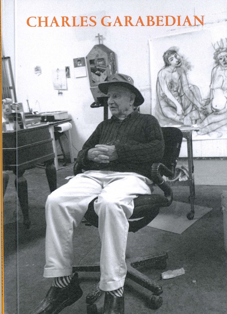 Black and white image of the artist reclining in an office hard in his studio. Art work hangs on the wall behind him.
